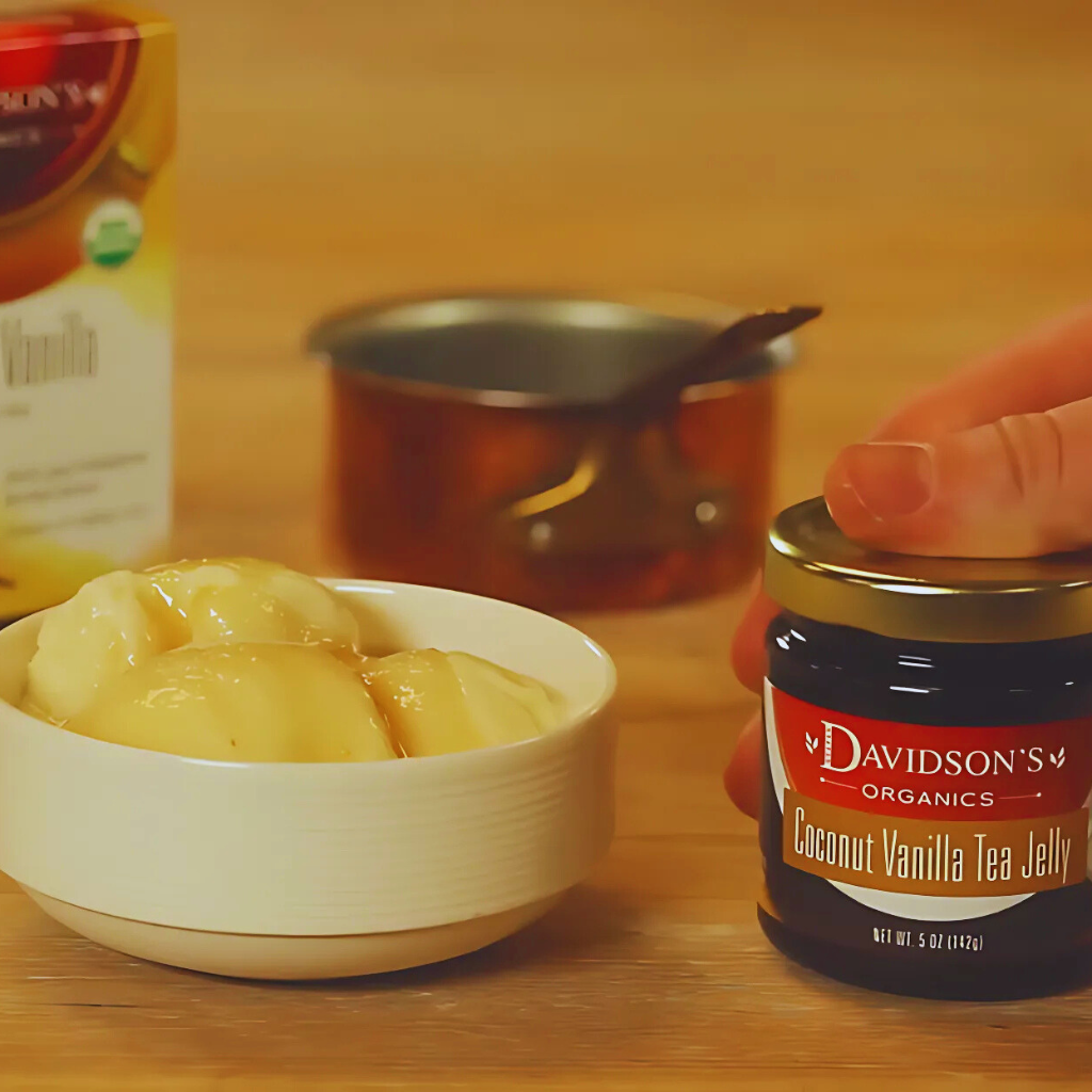 Jar of Davidson's Organics Coconut Vanilla Tea Jelly on a wooden table with a bowl of jelly and a carton in the background.