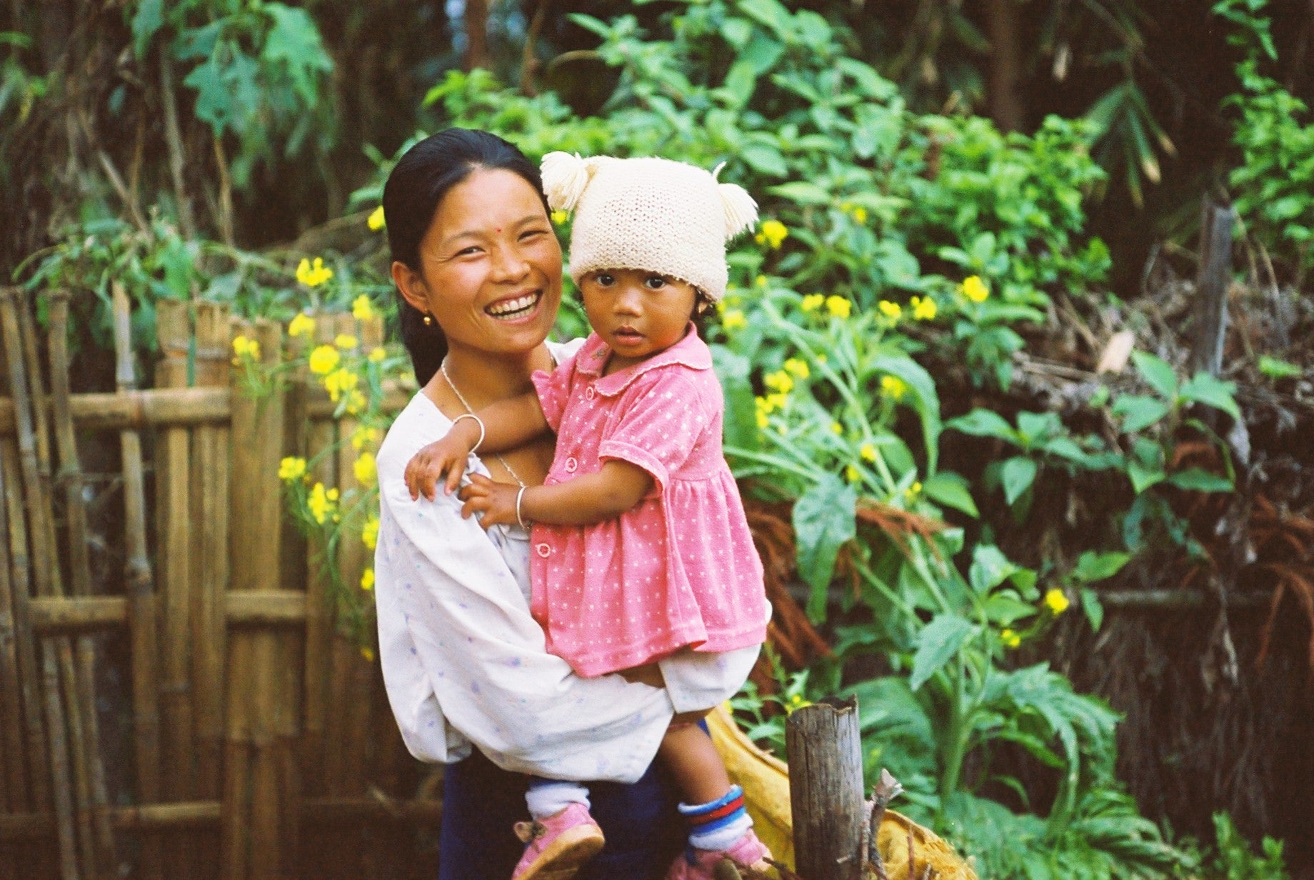 A smiling woman holding a young child in a white bear hat, standing in a lush garden.