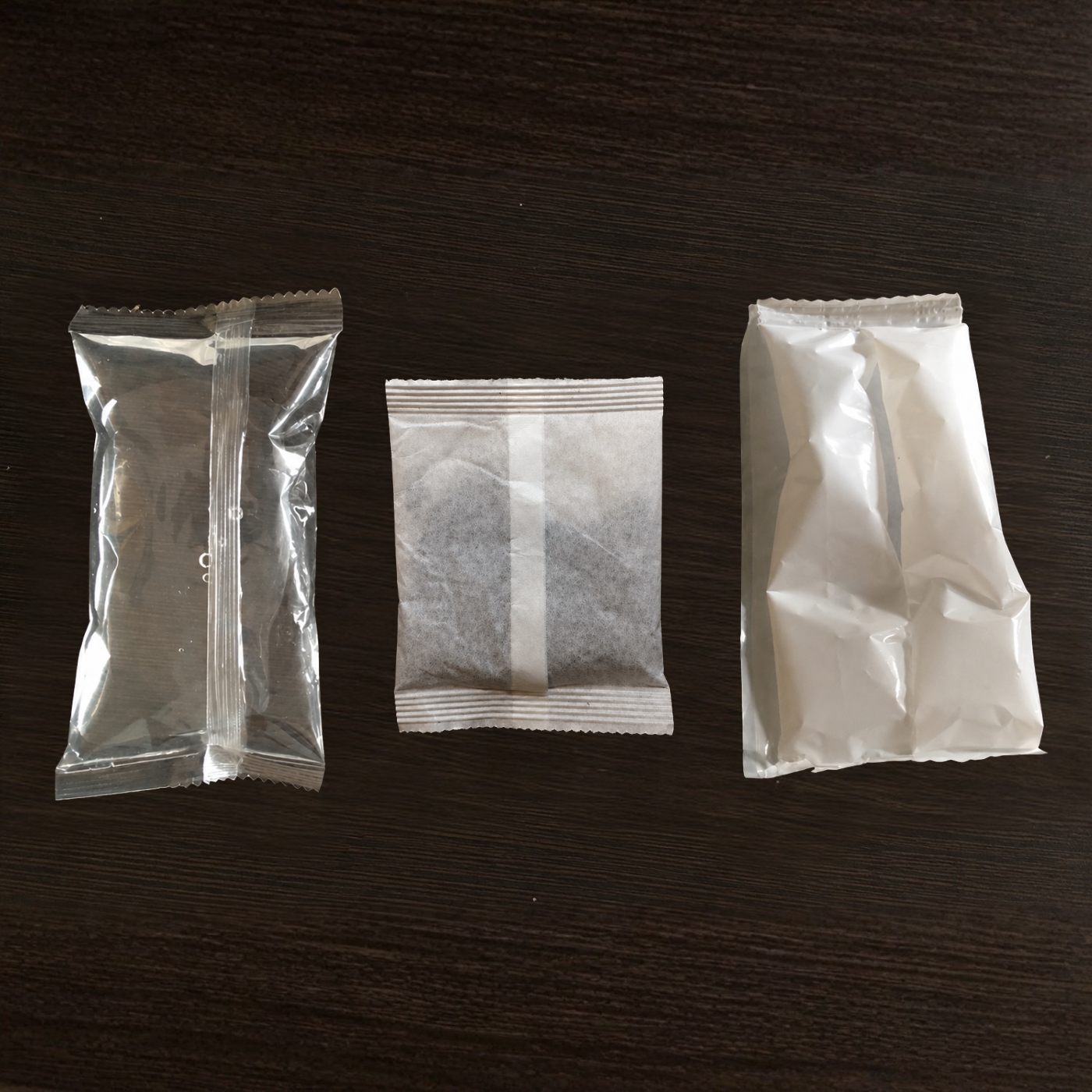 Three small sealed packets arranged side by side on a dark brown wood surface: a clear plastic packet on the left, a white paper packet with a vertical white stripe in the center, and a white paper packet on the right.