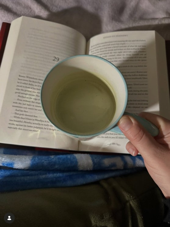 A hand holding a light blue and white ceramic mug with a pale greenish liquid inside, positioned over an open book on a bed with a blue blanket.