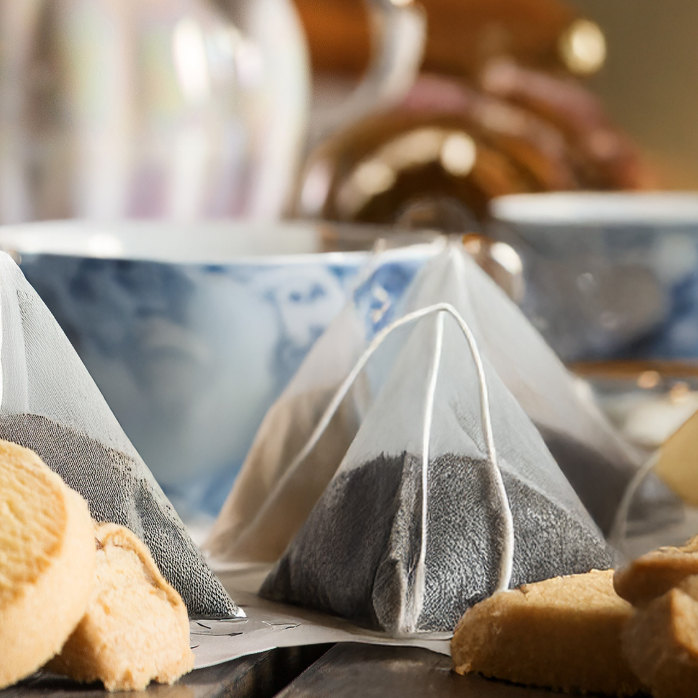 Tea bags in pyramid shape with a blurred background of tea cups and cookies.