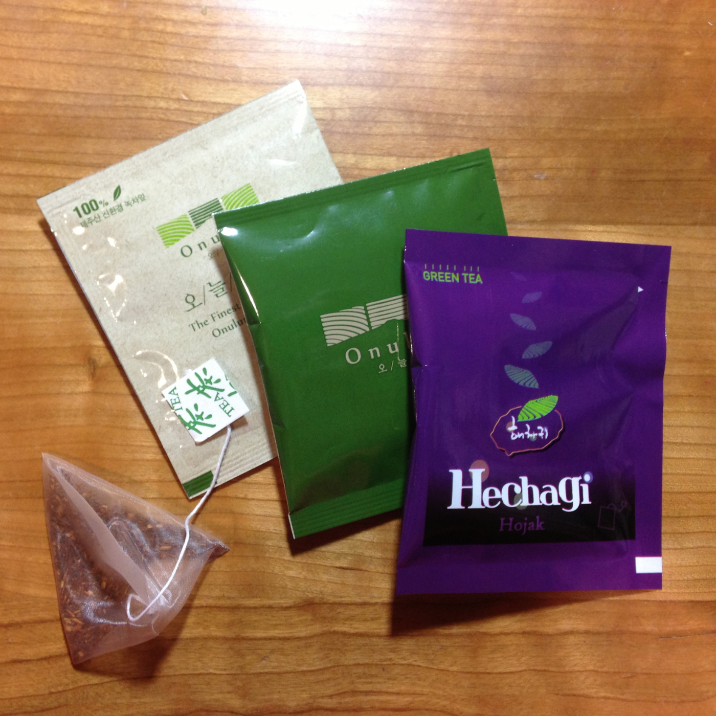 Three tea bags from Onui and Hechagi displayed on a wooden table, including a green tea bag, a purple Hechagi Hojak tea bag, a beige Onui tea bag, and a small triangular tea bag with its tag visible.