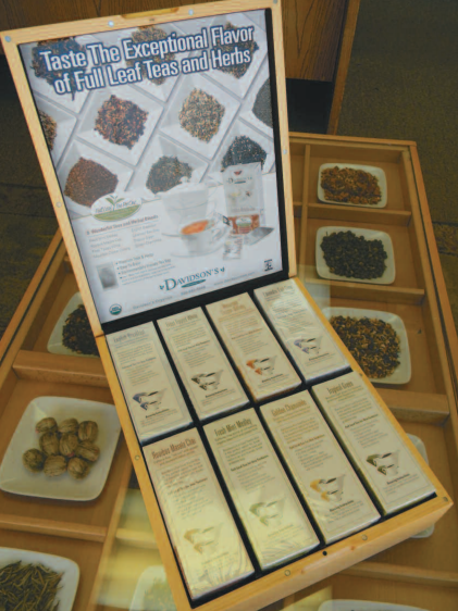 An open wooden display box filled with individually wrapped tea samples and a large printed tea information panel behind it, photographed indoors on a display table.