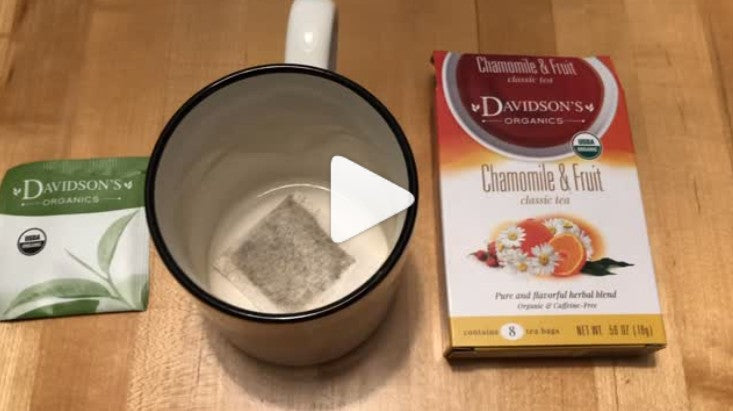 Davidson's Organics Chamomile & Fruit herbal tea shown with its retail box, a single tea bag, and an empty ceramic mug on a wooden tabletop.