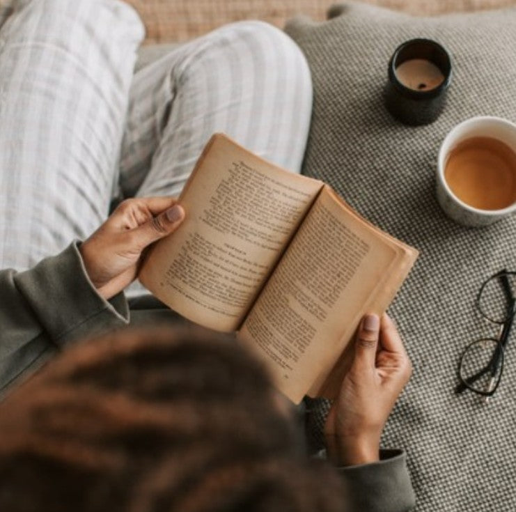 A person sitting on a couch reading an open book, with a cup of tea, a candle, and eyeglasses nearby in a cozy indoor setting.