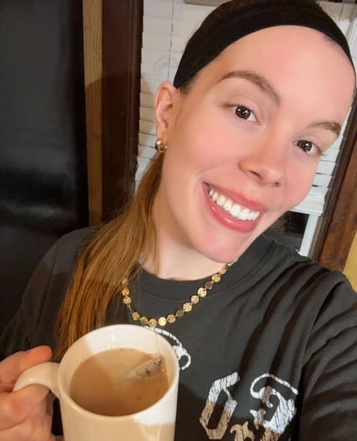 A woman smiling for a selfie while holding a white mug filled with a tea bag. She is wearing a black headband, a black graphic t-shirt, and a gold beaded necklace.