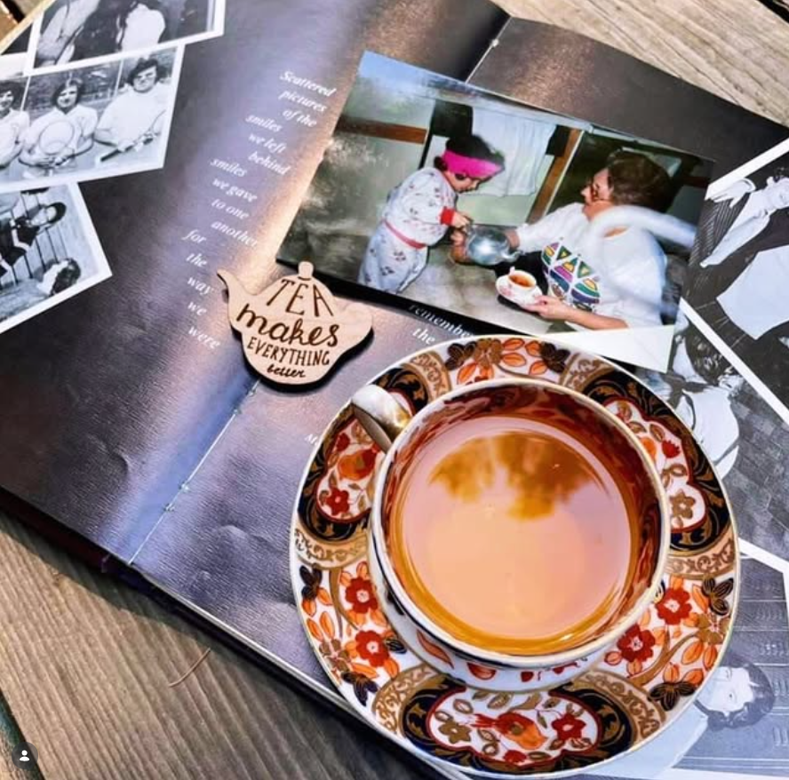 A close-up of a ceramic teacup and saucer filled with tea, placed on top of an open book surrounded by printed photos and a small wooden tag that reads 'tea makes everything better.'.
